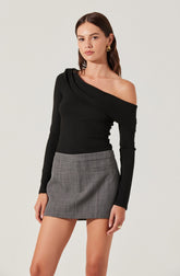 A woman is wearing a black, one-shoulder long-sleeve top with a draped detail on the shoulder, paired with a high-waisted, gray plaid mini skirt.